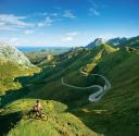 A cyclist on a mountaintop observes a landscape of mountains with the sea in the background