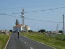 A cyclist rides along a road that runs in front of a coastal lighthouse