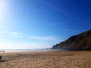 A wide beach with golden sand, cliffs, the sea in the background, and an intense blue sky