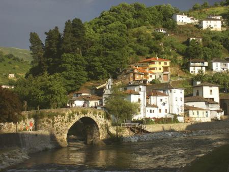 A single-arch stone bridge crosses a river next to a village of white houses on a hillside