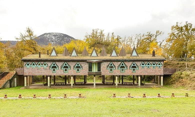 A building with unique architecture stands on pillars in a clearing, below between the pillars is the rest area. The building has a sloping roof with triangular windows simulating trees and is surrounded by trees with autumn foliage. In the background is a snow-capped mountain.
