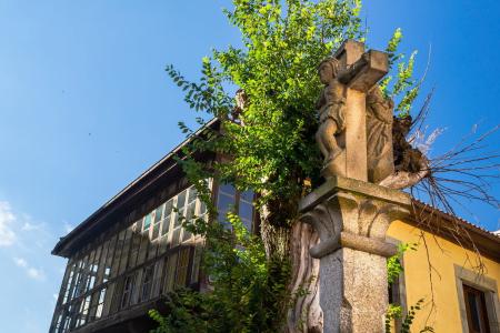 A stone wayside cross in the foreground, with a wooden gallery of a house and a tree behind