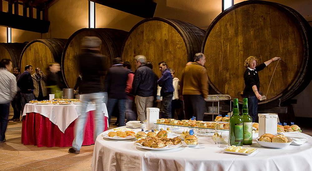 Espicha in a cider house with the snack tables in the foreground and the people and the cider barrels in the background.