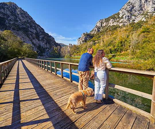 Couple sur une passerelle en bois avec leur chien observant le réservoir de Valdemurio.