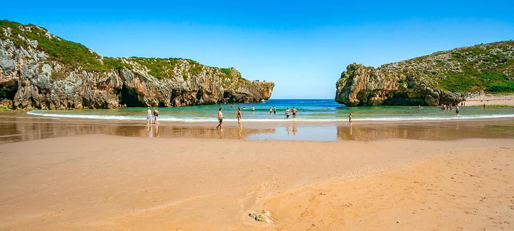 Vista panorámica de la Playa de Cuevas del Mar en Llanes, una impresionante cala con cuevas y arcos rocosos, bañistas y aguas cristalinas.