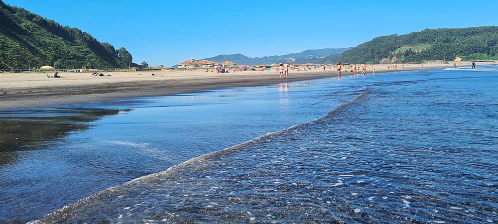 Una ola rompiendo en la orilla de la playa de los Quebrantos con personas caminando y disfrutando del día en la arena. Montañas al fondo.