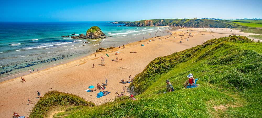Vista panorámica de la Playa de Penarronda en Castropol, Asturias, con su característico islote rocoso, arena dorada y bañistas disfrutando del sol.