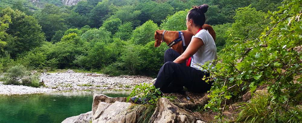 Vista de una mujer y su perro disfrutando de la naturaleza en la Olla de San Vicente, con el río Dobra y su entorno montañoso de fondo.