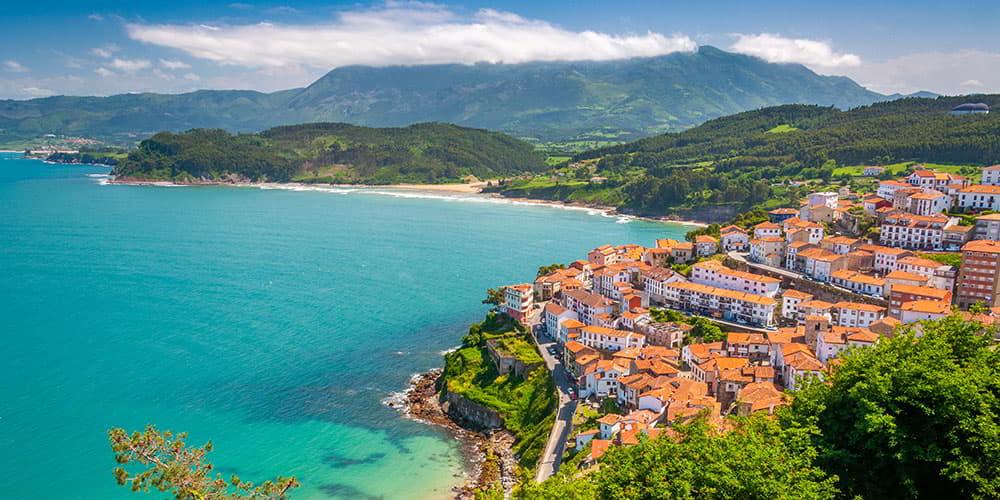 Vue panoramique de Lastres depuis le point de vue de San Roque, avec le hameau en terrasses sur la pente descendant vers la mer Cantabrique, les toits rougeâtres, le littoral et la mer bleue en arrière-plan par une journée ensoleillée.