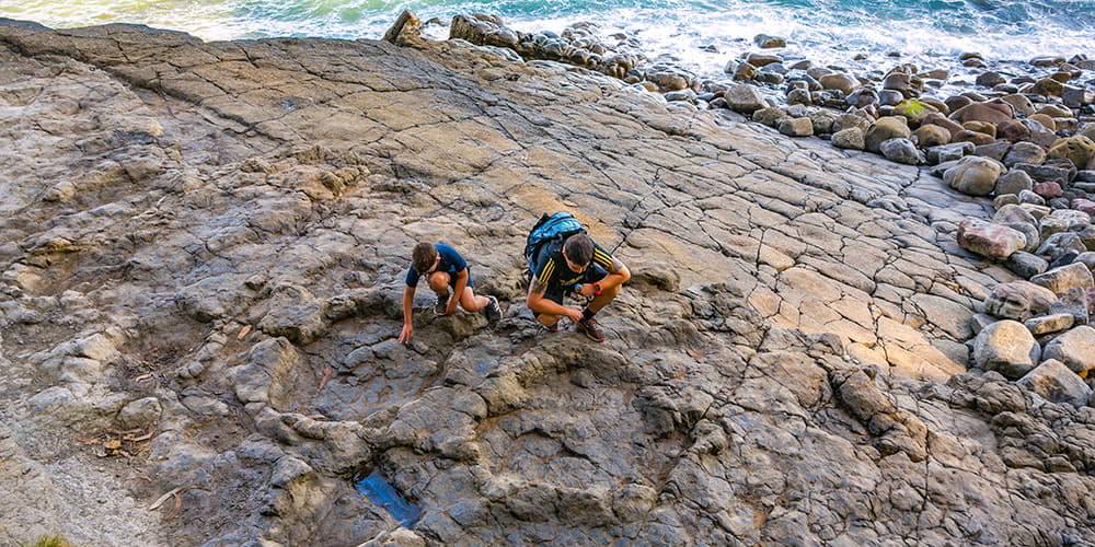Deux enfants s'accroupissent sur la roche pour observer les empreintes fossiles du gisement d'ichnite sur la plage de La Griega (Colunga) ; à l'arrière-plan, on aperçoit une partie de la mer dans une vue plongeante sans horizon défini.