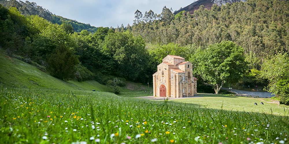 Panoramic view of San Miguel de Lillo, with the church standing out in the middle of a field of tall grass with wild flowers and trees in the background.