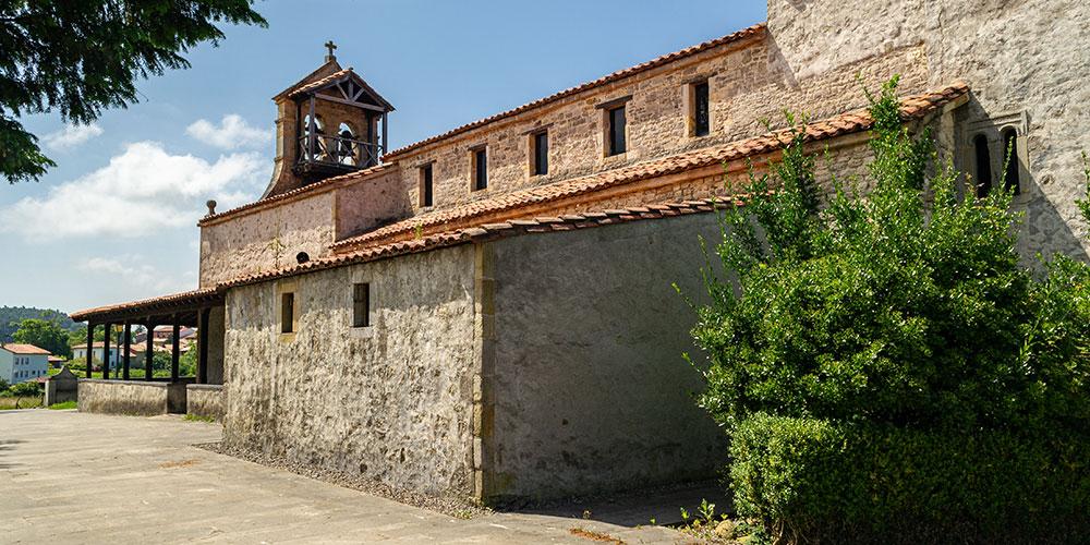 Façade arrière en pierre de l'église préromane de Santiago de Gobiendes, avec en arrière-plan un clocher à deux travées et des bâtiments traditionnels de Gobiendes.