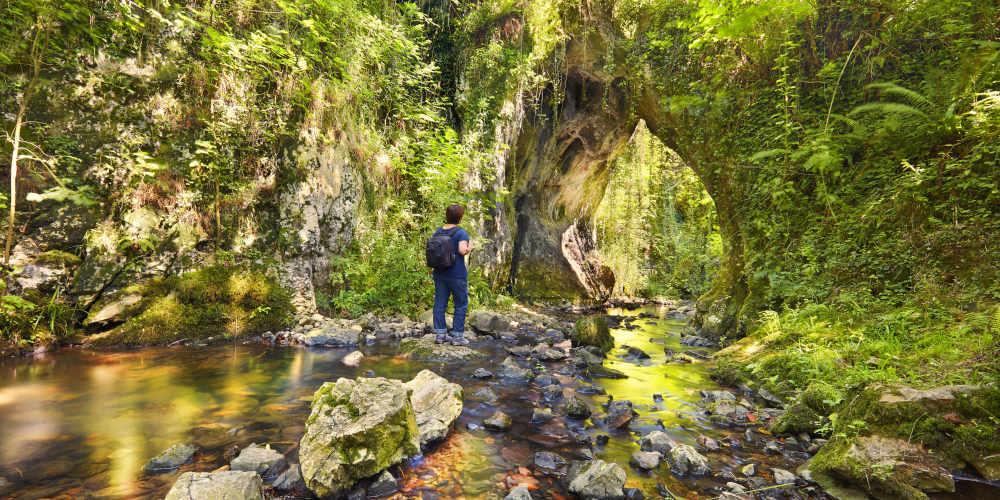 Woman on her back on a rock in the river Tuernes, in Los Covarones de Tuernes (Llanera), dressed in jeans, navy blue T-shirt and black rucksack; she stands in front of a natural arch of limestone rock covered with vegetation, with the stream flowing below and riparian vegetation all around.