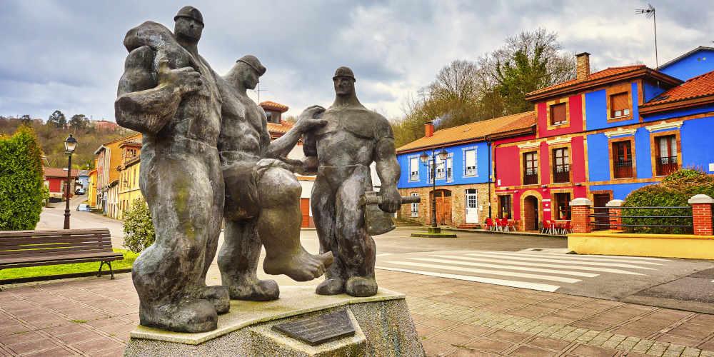 Monument to the miner in Carbayín Bajo (Siero): three massive bronze figures representing miners; one holds an axe in his left hand, another carries a bundle over his shoulder and the central figure leans on his companions while raising his right leg in an attitude of effort. The sculptures, of a sturdy 'Hulk' or 'Stone Man' appearance, stand on a stone pedestal. In the background, behind a pedestrian crossing, bright maroon and blue houses stand out in an open urban environment.