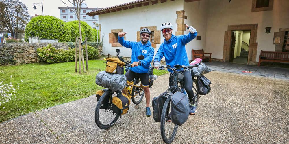 Two pilgrims on bicycles on the Camino de Santiago, equipped with backpacks and packs strapped to the bikes at the front and back; both wearing sky blue mackintoshes with a flag similar to the Argentinean flag, wearing cycling glasses and posing with smiling thumbs up, next to a cobbled passage with a small garden and a tree next to it.