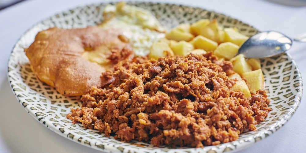 Close-up of a plate of picadillo with tortos and potatoes, served on a plate decorated with a mosaic motif on a white tablecloth, accompanied by a spoon resting on the rim.