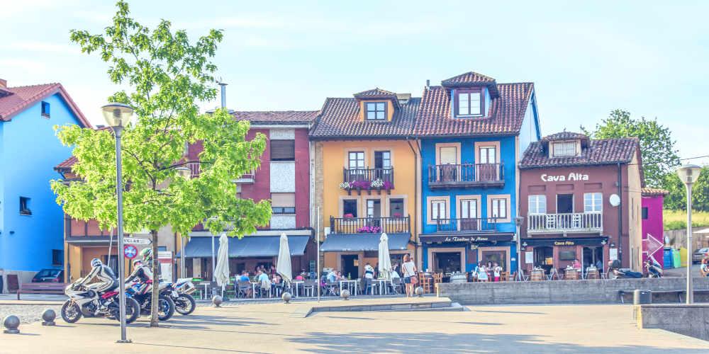 Plaza Les Campes in Siero, with houses with facades painted in bright yellow, blue and brown; under the arcades there are terraces with people having a drink, while in the square there is a tree and several motorbikes parked next to the cleared paved area.