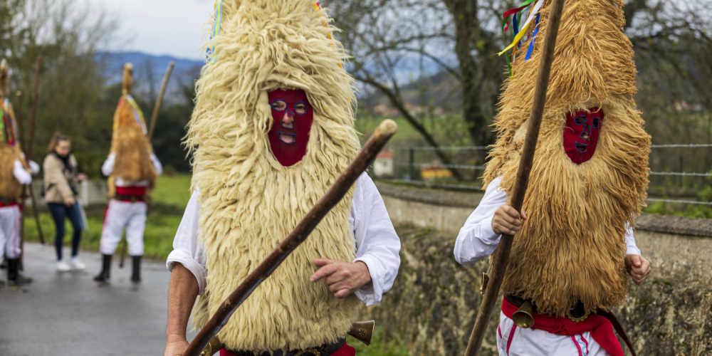 Two Sidros in the foreground during the Valdesoto winter masquerade, wearing the traditional costume consisting of a white shirt and trousers, a sash and coloured ribbons, a mask and a long stick in their hands; in the background, blurred, two other Sidros can be seen with their backs turned.
