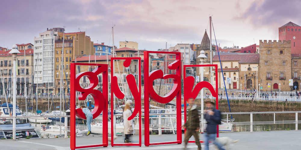 Rotes Schild der Letronas von Gijón mit Passanten im Vordergrund; dahinter der Jachthafen mit Booten und Segelbooten, Gebäude im Zentrum, der Uhrenturm und der Palast von Revillagigedo.