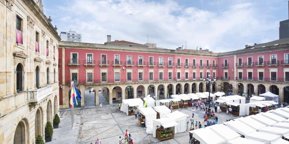 Luftaufnahme der Plaza Mayor in Gijón während des Kunsthandwerker- und Ökomarktes, aufgenommen von einem hohen Fenster aus; das Rathausgebäude ist von der Seite zu sehen und im Vordergrund die Flaggen Spaniens und der Europäischen Union an den Masten vor dem Gebäude.
