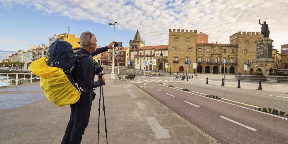 Pilger auf der Promenade des Jachthafens in Gijón, der mit seinem Handy ein Foto in Richtung der Pelayo-Statue und des Palastes von Revillagigedo macht; er trägt einen Wanderrucksack und hält Wanderstöcke in der Hand.