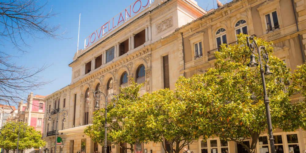 Fassade des Theaters Jovellanos in Gijón an einem sonnigen Tag, mit blauem Himmel und Bäumen am Paseo de Begoña im Vordergrund.