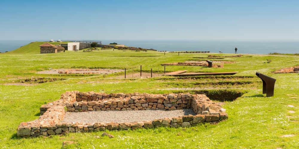 Gesamtansicht des archäologischen Parks Campa Torres in Gijón mit Resten von Verteidigungsanlagen und Wohnhäusern in einer Küstenlandschaft.