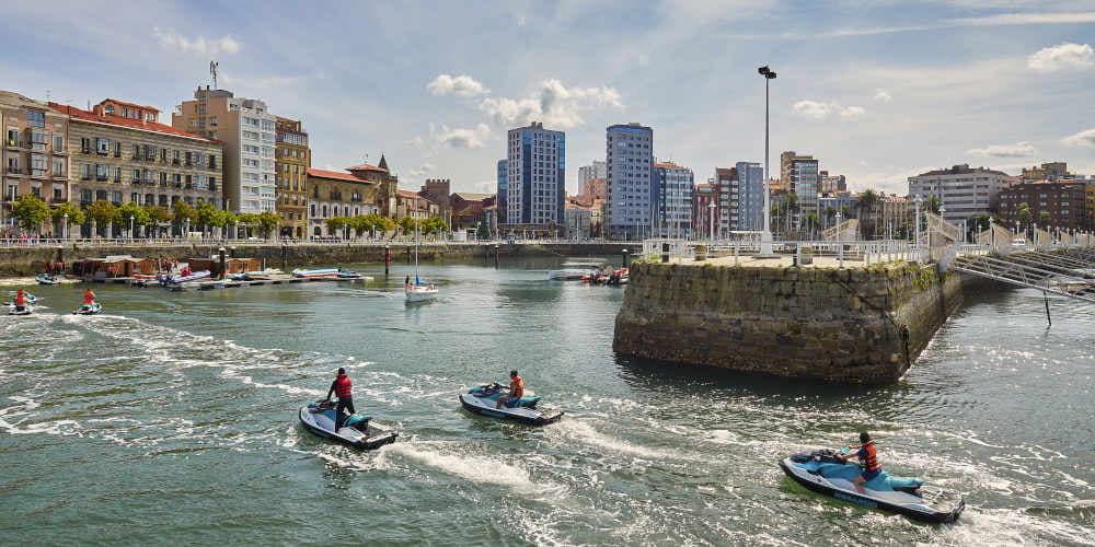 Jetski-Verleih im Jachthafen von Gijón, mit drei Personen, die auf ihren Jetskis zum Kai zurückkehren; im Hintergrund sind die Hafenpromenade, das Denkmal der Apfelweinflaschen und mehrere städtische Gebäude zu sehen.