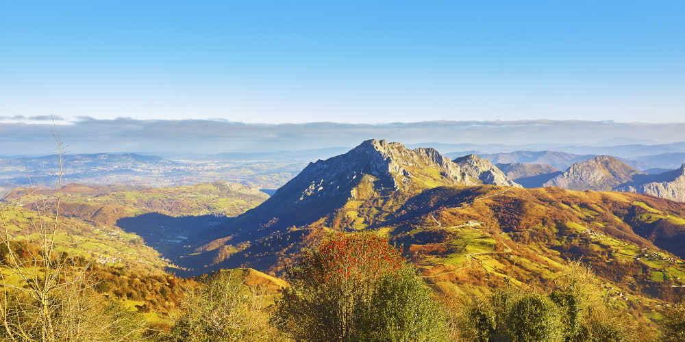 Vista panorámica del Monsacro desde la subida al Angliru, en Riosa, con laderas en tonos verdes y anaranjados; al fondo se distinguen áreas rurales y parte del entorno urbano del área central asturiana.