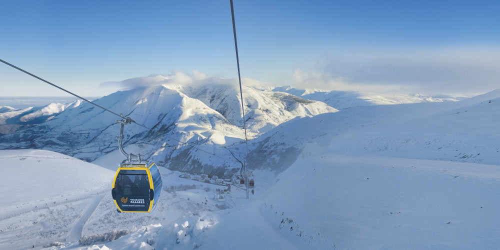 Telesilla amarillo en funcionamiento con personas en su interior en la Estación Invernal y de Montaña Valgrande-Pajares, rodeado de un paisaje de montaña completamente nevada.