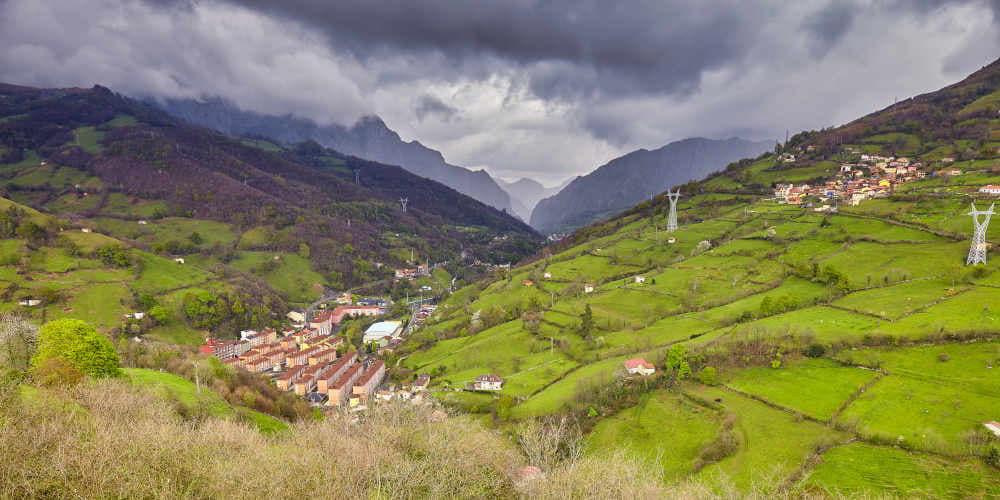 Vista de L’Arra desde el Mirador del Aramo, en Riosa, bajo un cielo nublado; en la panorámica se distinguen zonas de prados y bosques, un núcleo de viviendas en el valle y otro asentamiento más elevado en la ladera derecha.