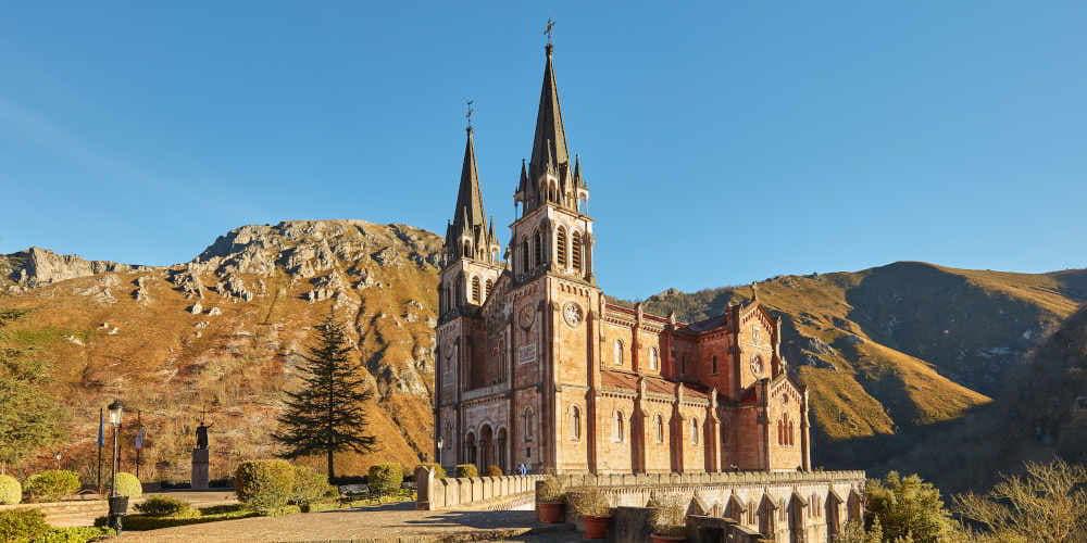 Vista del Real Sitio de Covadonga desde el lateral derecho, con barandilla de piedra y macetas en primer plano; al fondo se alza la basílica y la montaña en tonos otoñales y áridos, menos verdes de lo habitual en el paisaje asturiano.