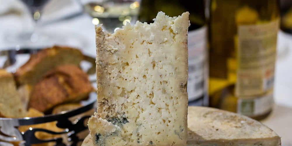 Primerísimo plano de un corte de queso Gamonéu que muestra su textura porosa, con el bloque completo difuminado al fondo; detrás se distinguen dos botellas de vino y un recipiente de acero inoxidable con pan rústico troceado, de aspecto similar al de maíz.