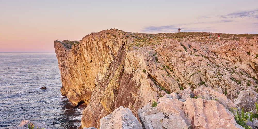 Acantilados de Pría con formaciones rocosas en primer plano y vista lateral del acantilado al fondo, donde varias personas caminan hacia el extremo y dos jóvenes se sitúan en la punta; a la izquierda se extiende el mar con el horizonte en tonos difuminados rosas y el cielo azul tenue de un atardecer.