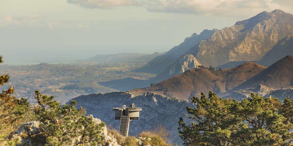 Vista aérea del Mirador del Fitu desde la parte trasera, con dos personas en la plataforma apreciando el paisaje; a la derecha destacan cuatro árboles al borde del mirador y al fondo se extiende la panorámica característica con montañas verdes y la costa en el horizonte.