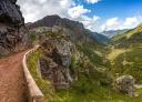 Dirt path on a sunny day with a wooden railing and views of a green and rocky valley.