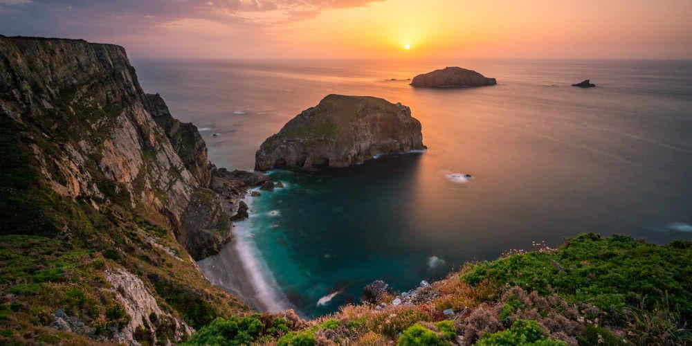 Atardecer sobre el Cabo Peñas visto desde lo alto, con tonalidades rojizas en el cielo y el mar. En el centro de la imagen destaca la Isla de La Erbosa, un islote rocoso que emerge frente al acantilado, en la costa del concejo de Gozón.