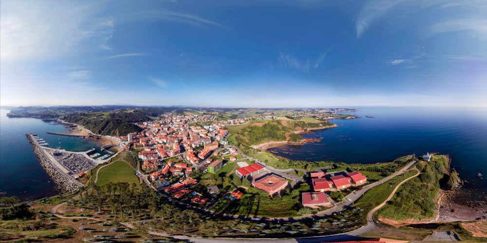 Vista aérea de Candás en formato panorámico, con mar y playas a ambos lados, núcleo urbano en el centro y cielo despejado.