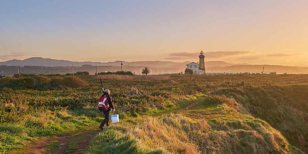 Vista del Faro de San Juan de Nieva al fondo, con un hombre caminando por un sendero entre vegetación, portando utensilios de pesca. La escena transcurre con luz cálida de amanecer o atardecer.