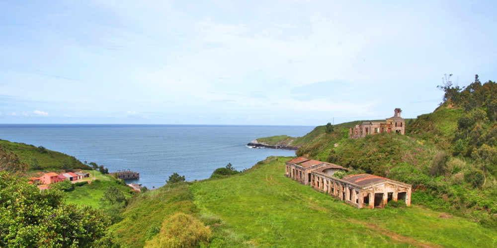 Vista del conjunto costero de las minas de Llumeres en Gozón, en estado de abandono entre vegetación. A la izquierda, se distinguen varias viviendas, en un entorno de prados verdes y costa acantilada.