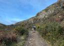 Two people walk along a rocky path in mountainous terrain.