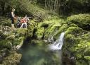 Wasserfall, umgeben von üppiger grüner Vegetation, mit einer am Wasser sitzenden Familie