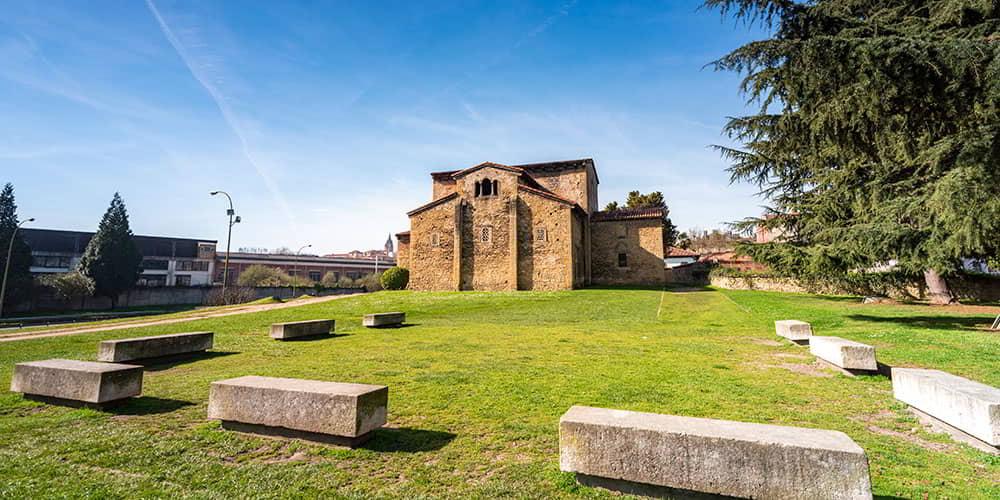 Rear view of the pre-Romanesque church of San Julián de los Prados in Oviedo/Uviéu, surrounded by a green lawn and stone blocks as seats.