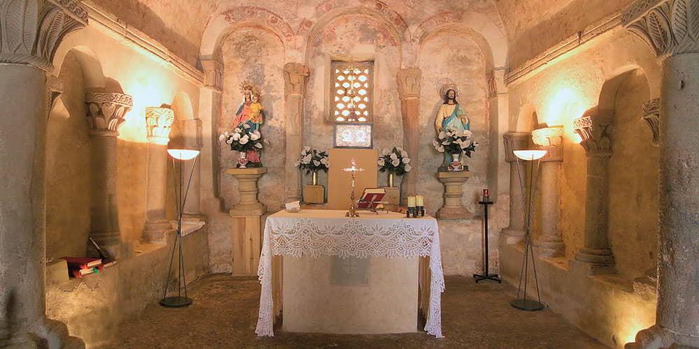 Ambiente interior de San Salvador de Priesca, destacando los arco, los capiteles decorados y las decoraciones murales, con un altar cubierto por un mantel blanco.