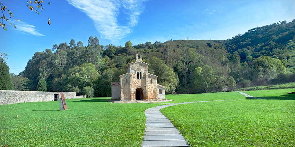 Exterior view of the church of San Salvador de Valdediós on a sunny day, with a path leading to its entrance.
