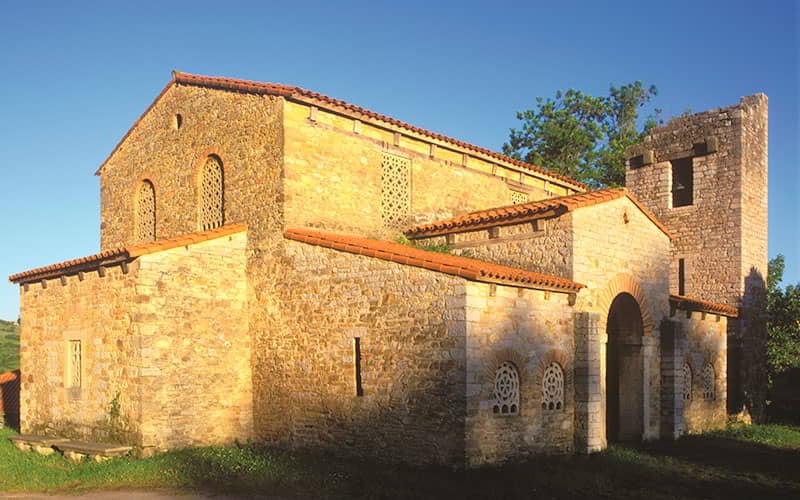 The pre-Romanesque church of Santa María de Bendones in Oviedo/Uviéu, showing the entire exterior and its latticed windows, tiled roofs and tower.