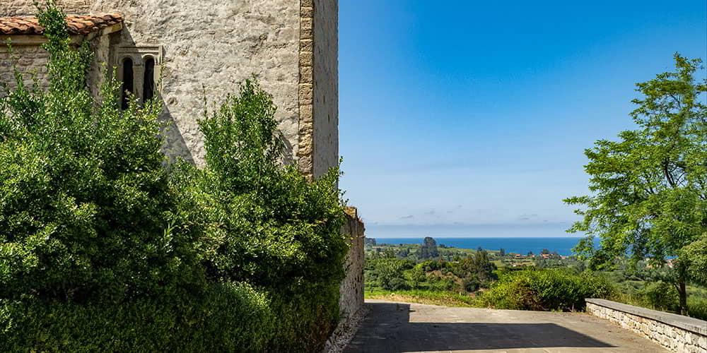A side of the church of Santiago de Gobiendes, showing a pre-Romanesque window and the Asturian landscape with the sea in the background.