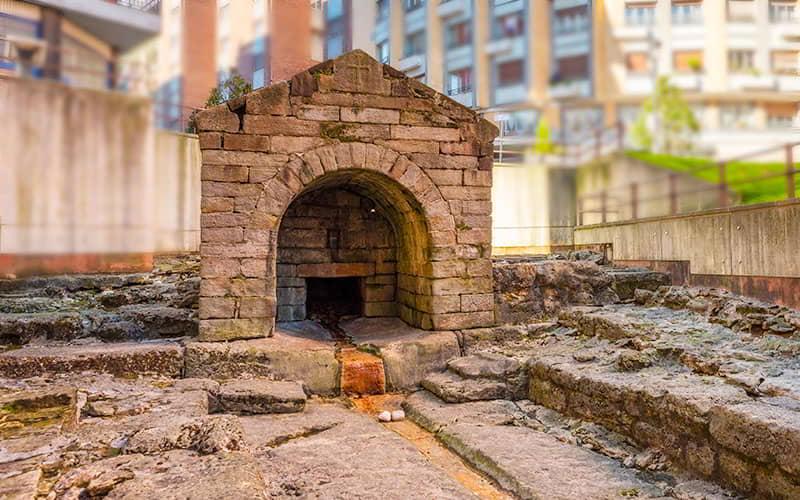 Front view of the Foncalada Fountain, a historic urban landmark, with the archaeological remains around it.
