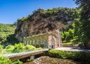 A stone bridge crosses a river to a stone building in a hilly and sunny landscape.