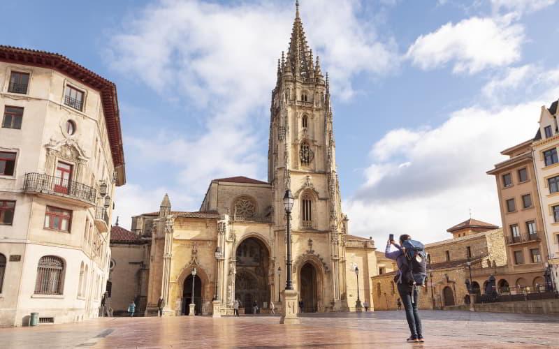 Peregrino en la plaza frente a la Catedral de San Salvador de Oviedo tomando una fotografía de la fachada con su teléfono móvil; lleva mochila con la concha del Camino de Santiago colgada.